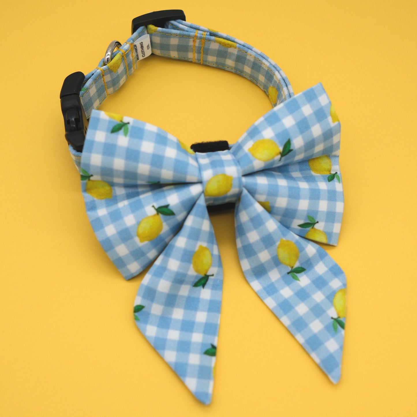 A light blue and white gingham pet collar with a matching large bow featuring a lemon print, displayed on a solid yellow background.