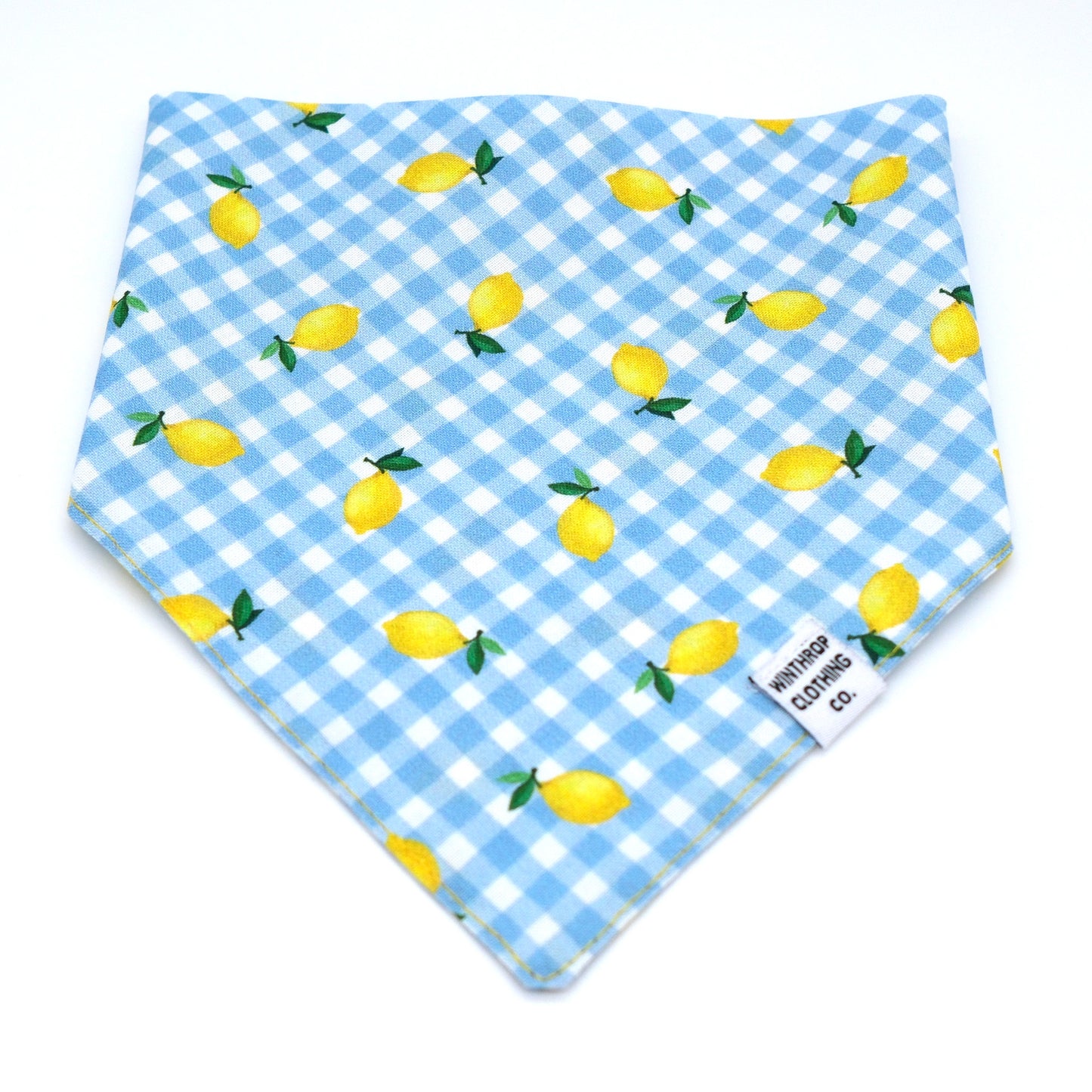 A triangular bandana bib featuring a light blue and white gingham check pattern with a scattered print of yellow lemons and green leaves. A small white fabric tag on the lower right corner reads 'winchester cotton co.